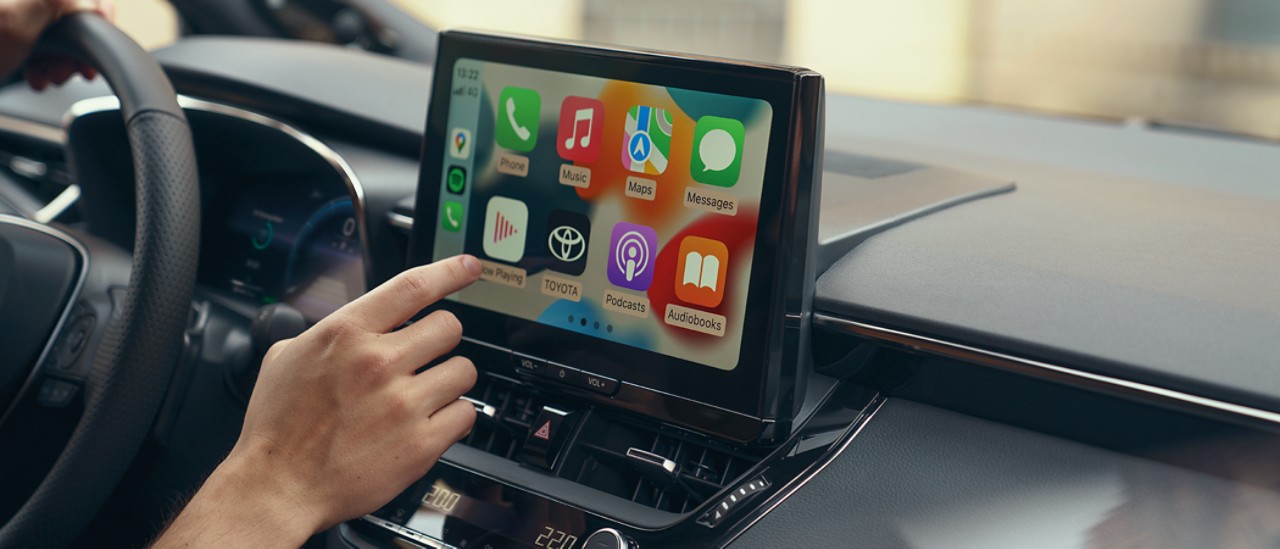 A person inside a Toyota interacts with the car's multimedia touch screen. The display shows the Apple CarPlay home screen.